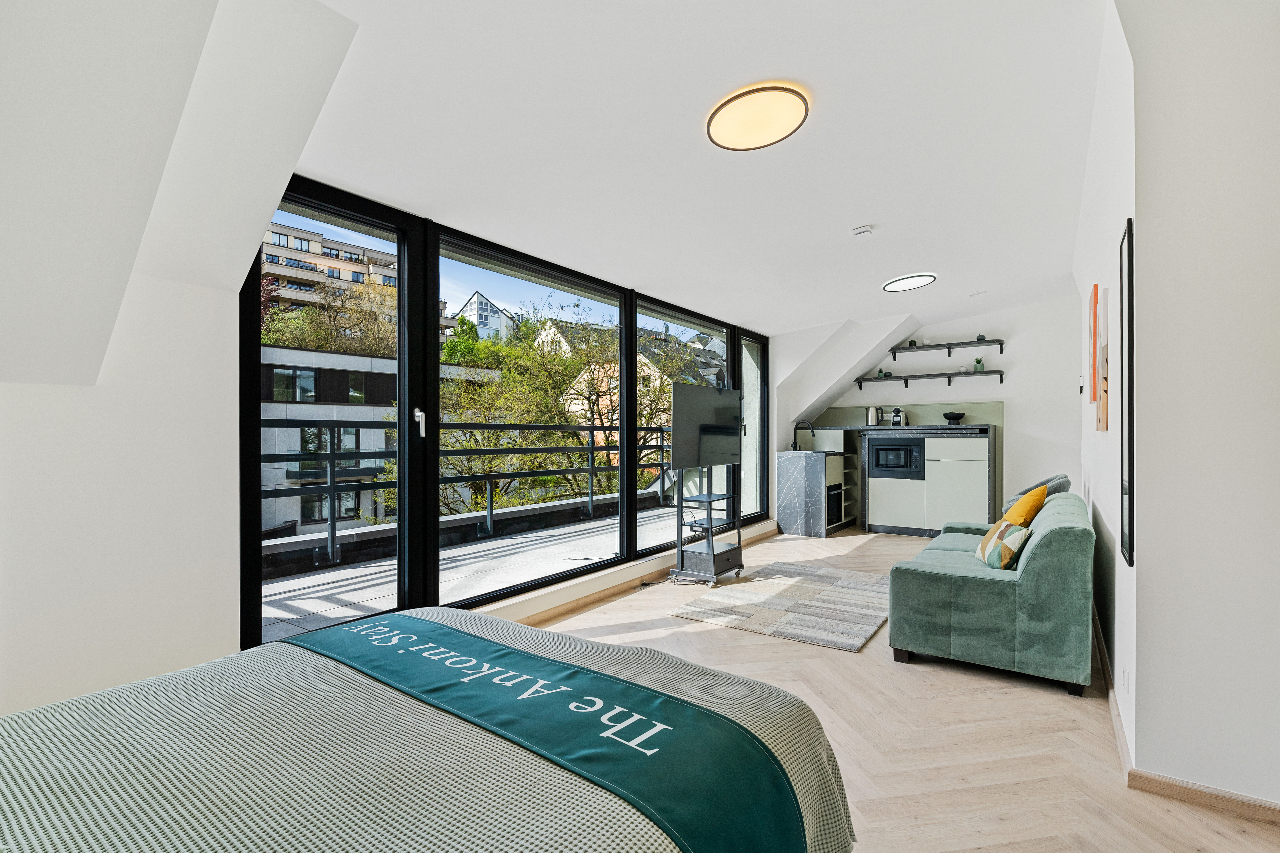 Bright studio apartment with a bed, green sofa, and kitchenette. Large windows offer a view of trees and buildings.