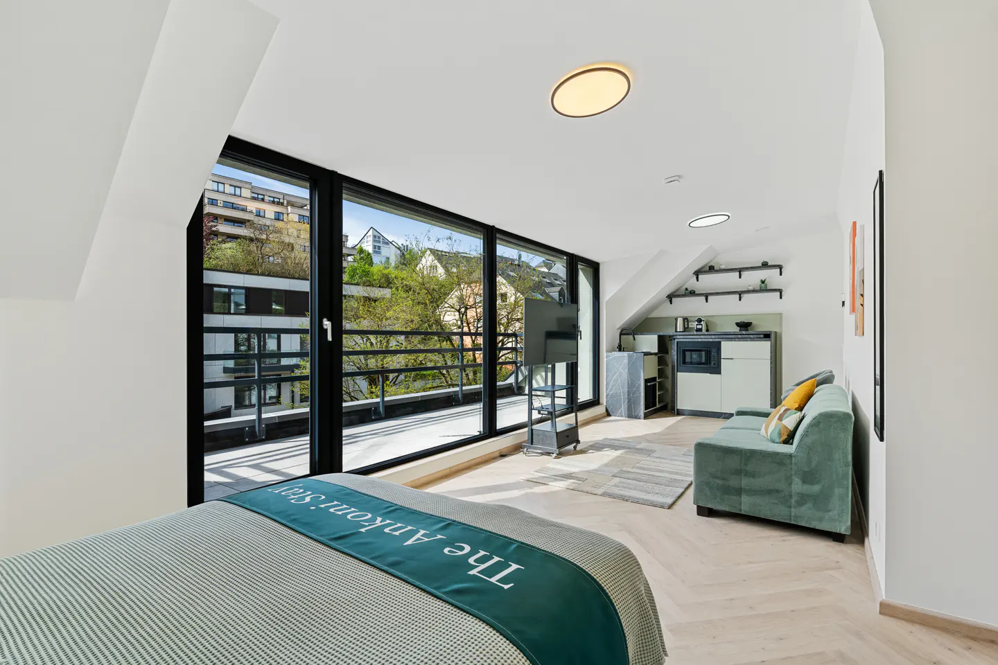 Bright studio apartment with a bed, green sofa, and kitchenette. Large windows offer a view of trees and buildings.