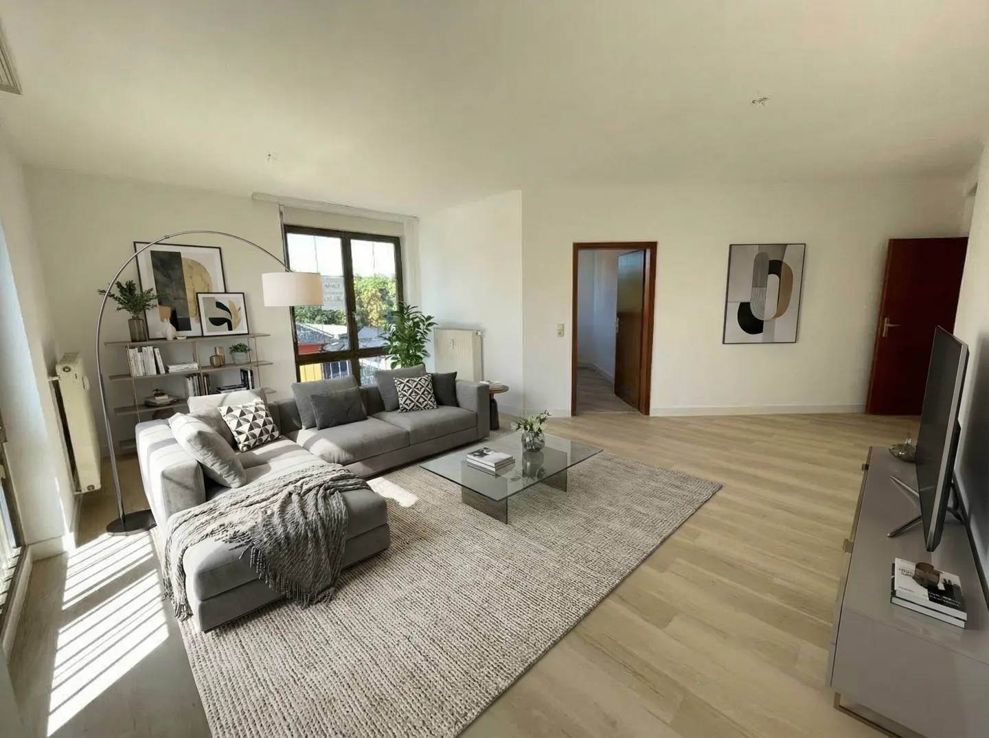 Bright living room with gray sectional sofa, glass coffee table on a textured rug, and light wood floors. Artwork and a TV are visible.