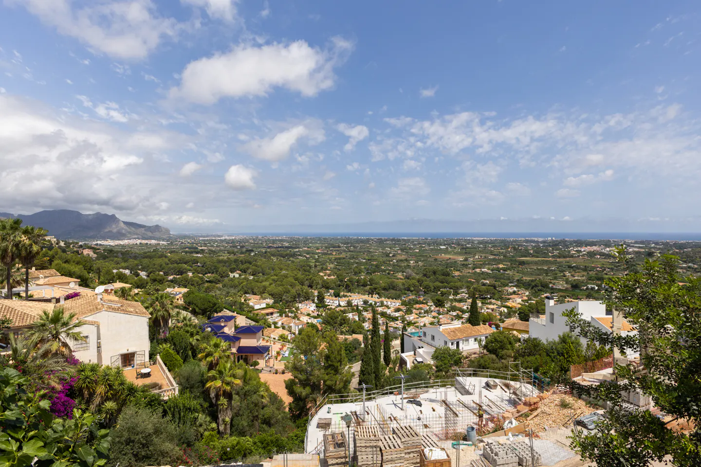 High-angle view of a Mediterranean hillside town with white villas, lush greenery, and a distant sea under a blue sky.