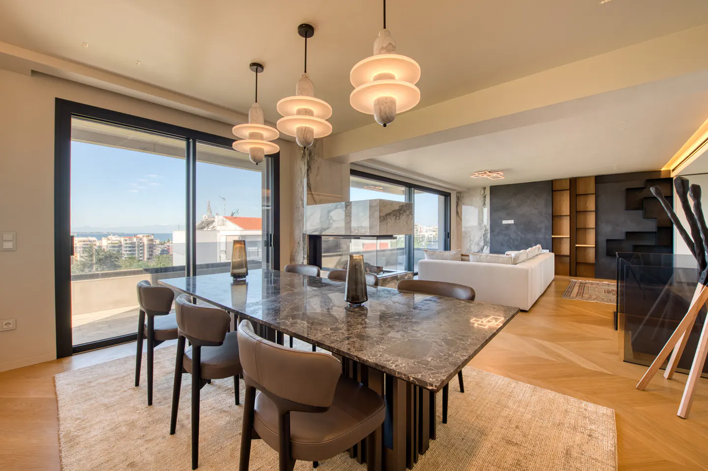 A modern dining room with a marble table, leather chairs, and unique pendant lights. Large windows offer a city view.