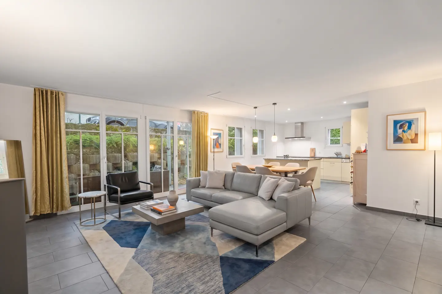 Open-concept living room with gray tile floors, a gray sectional sofa, and a geometric rug. Large windows with gold curtains overlook a garden.