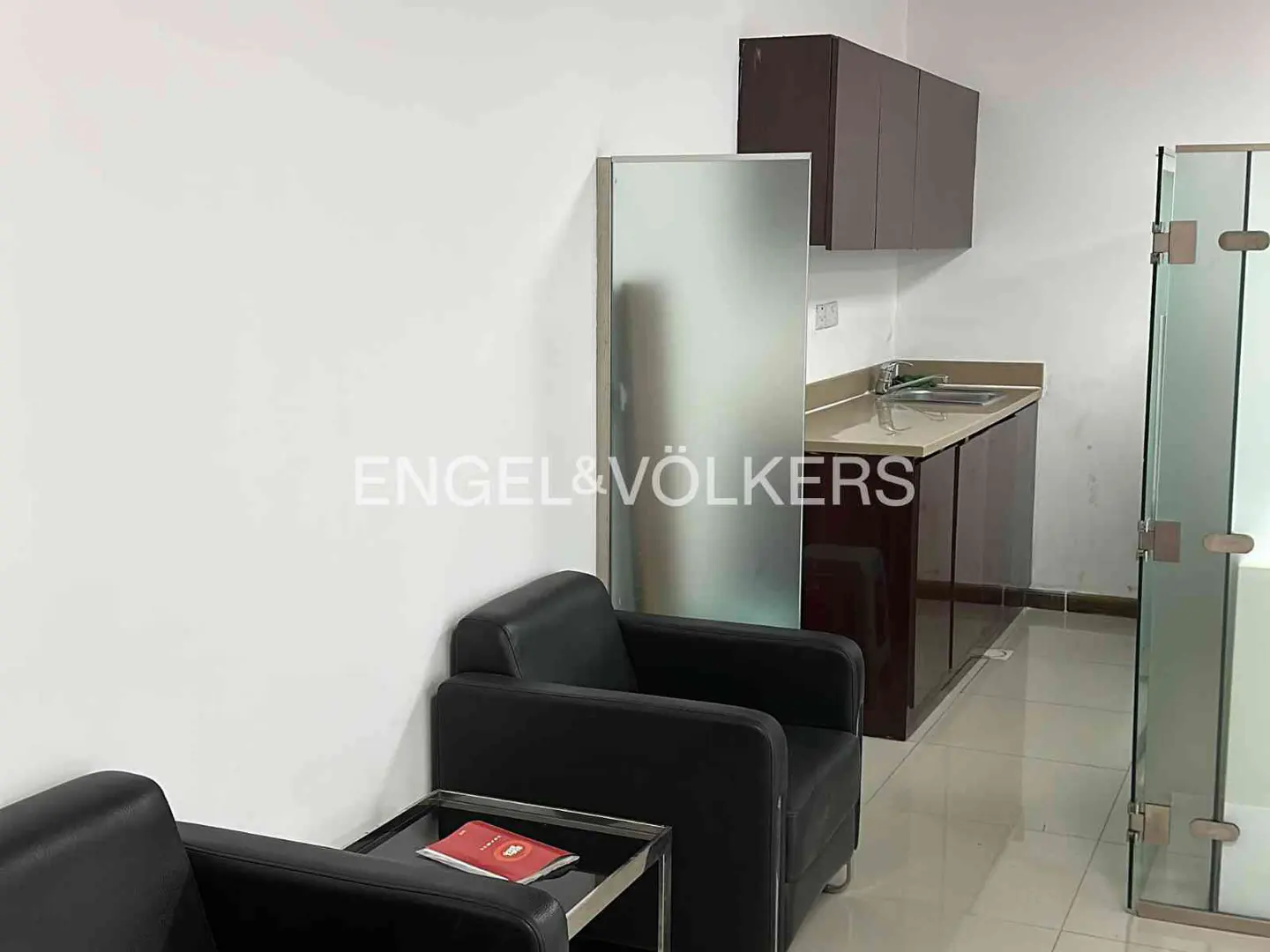Interior view of a real estate office with black armchairs, a small kitchen area, and a glass-walled room.