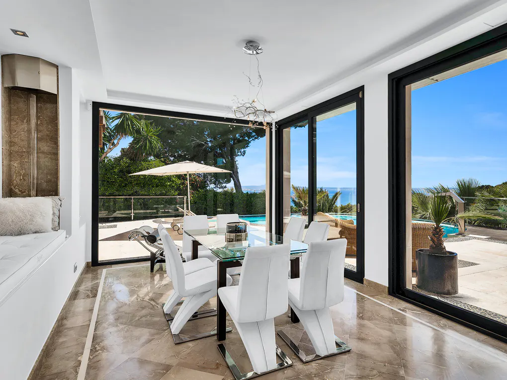 Bright dining room with marble floors, a glass table, and white chairs. Large windows offer a view of a pool, ocean, and blue sky.