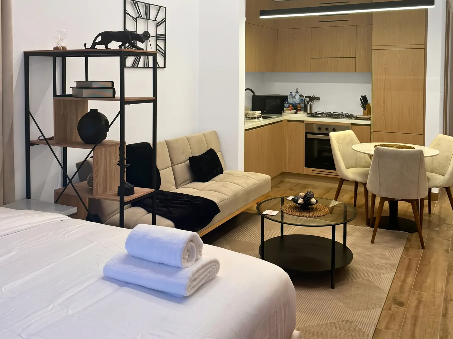 Modern apartment interior with a bed, sofa, kitchen, and dining area. White linens, beige sofa, and light wood cabinets create a bright, inviting space.