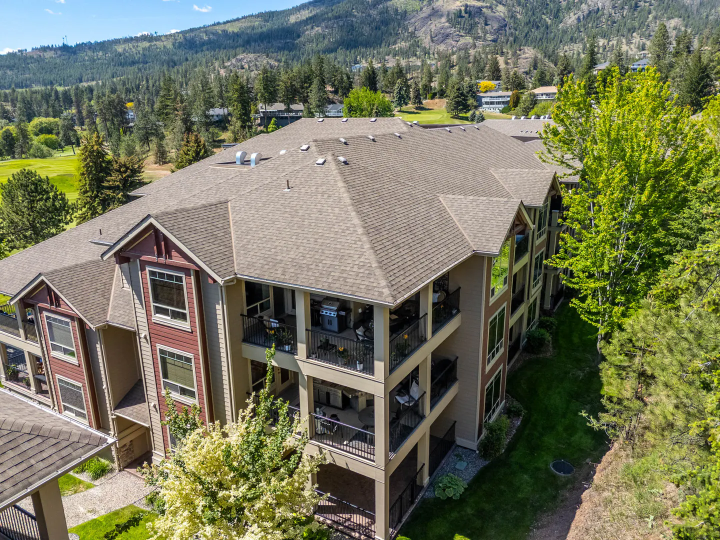 Aerial view of a three-story condo building with balconies, a brown roof, and a backdrop of green trees and mountains.