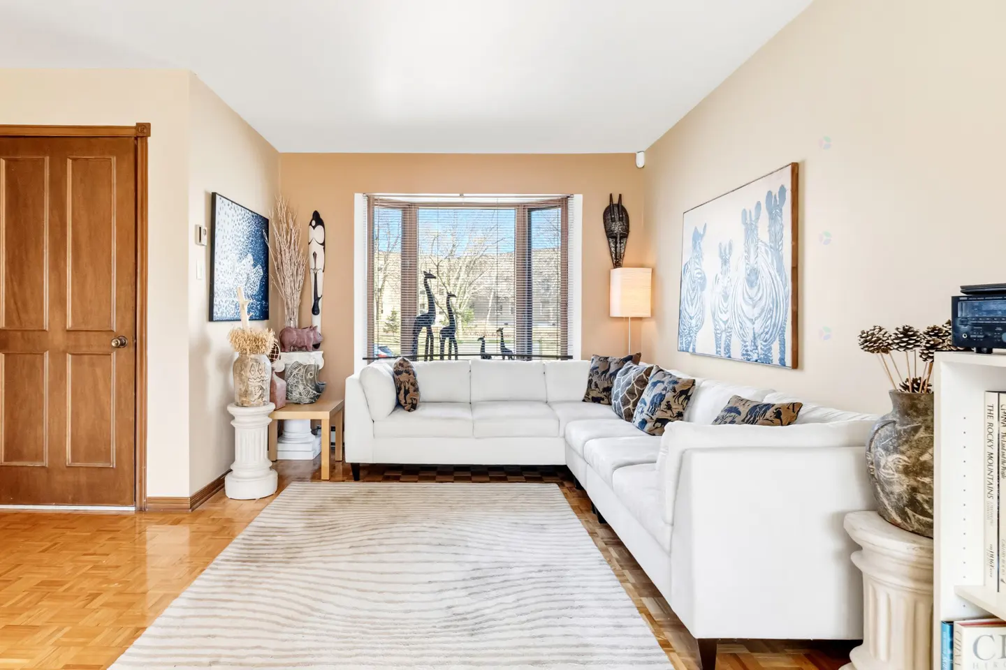 Living room with a white sectional sofa, patterned rug, and decor. A bay window overlooks trees. Artwork and African-inspired accents add character.