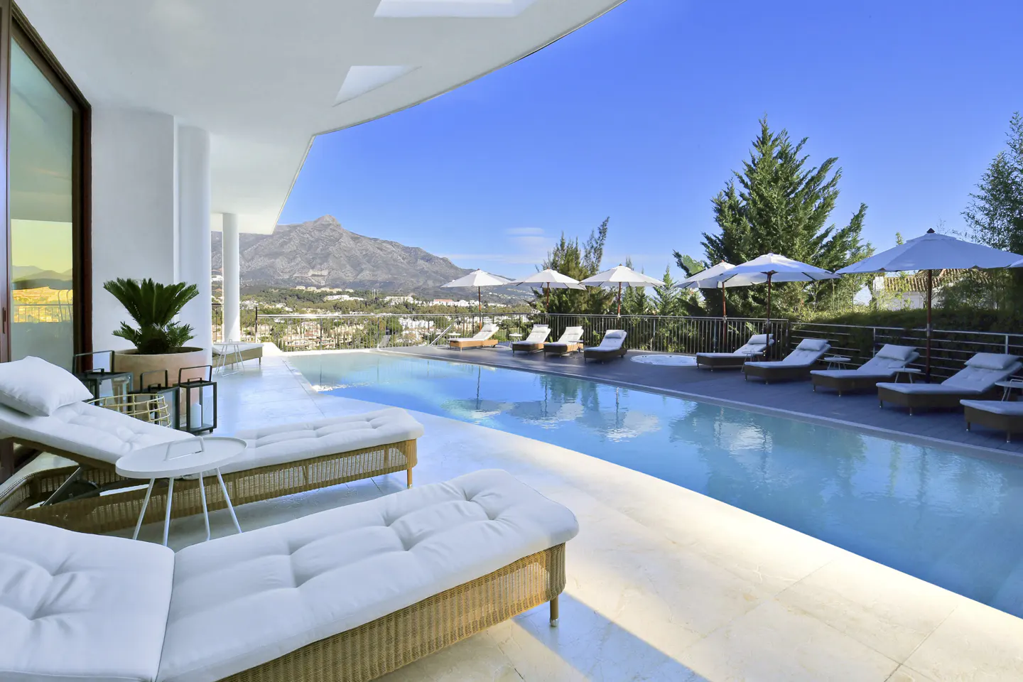 Luxury villa with a pool, white lounge chairs, and umbrellas. Mountain views and blue sky in the background.