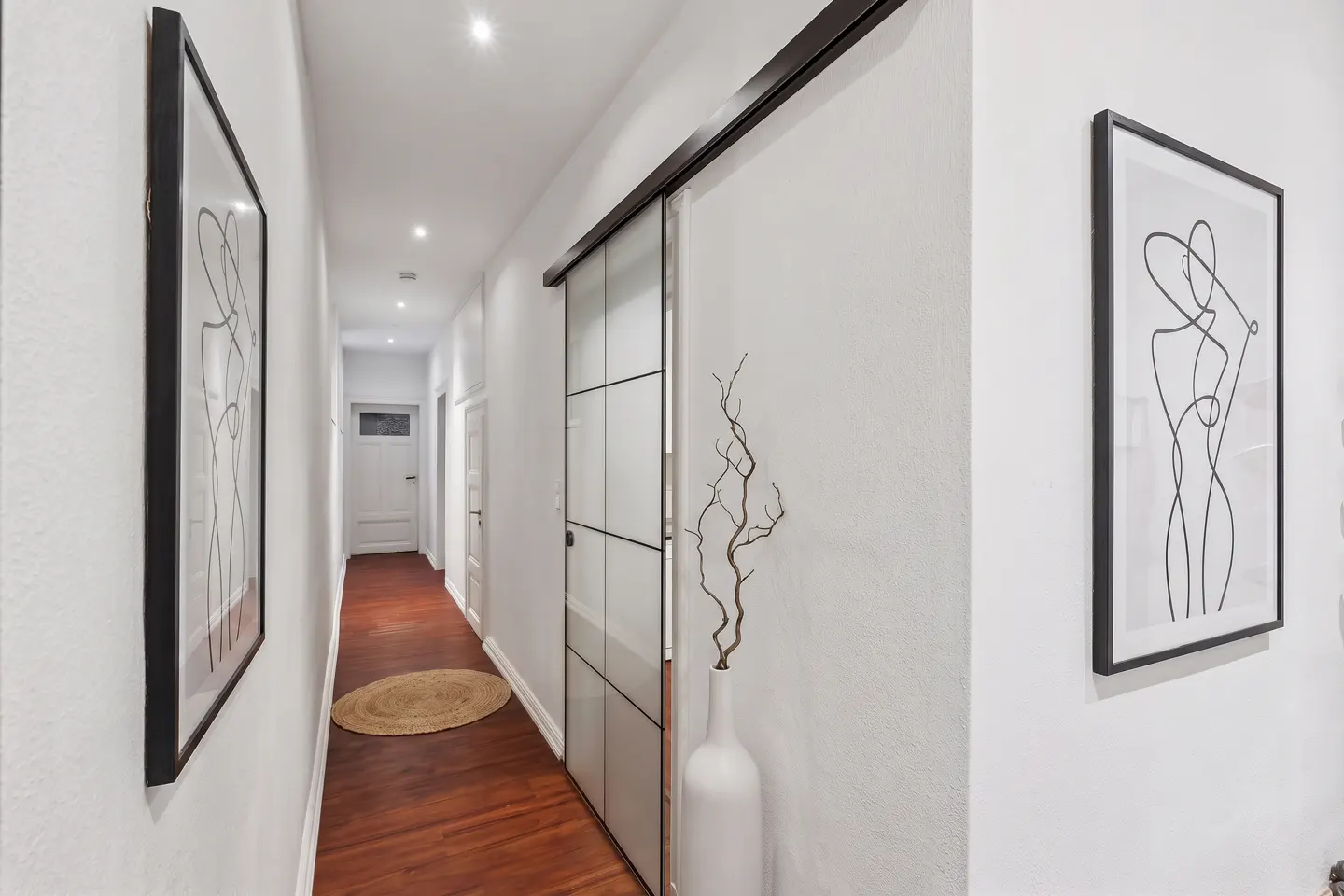Hallway with white walls, wood floors, and recessed lighting. Framed art hangs on the walls. A sliding glass door and a white vase with branches are visible.