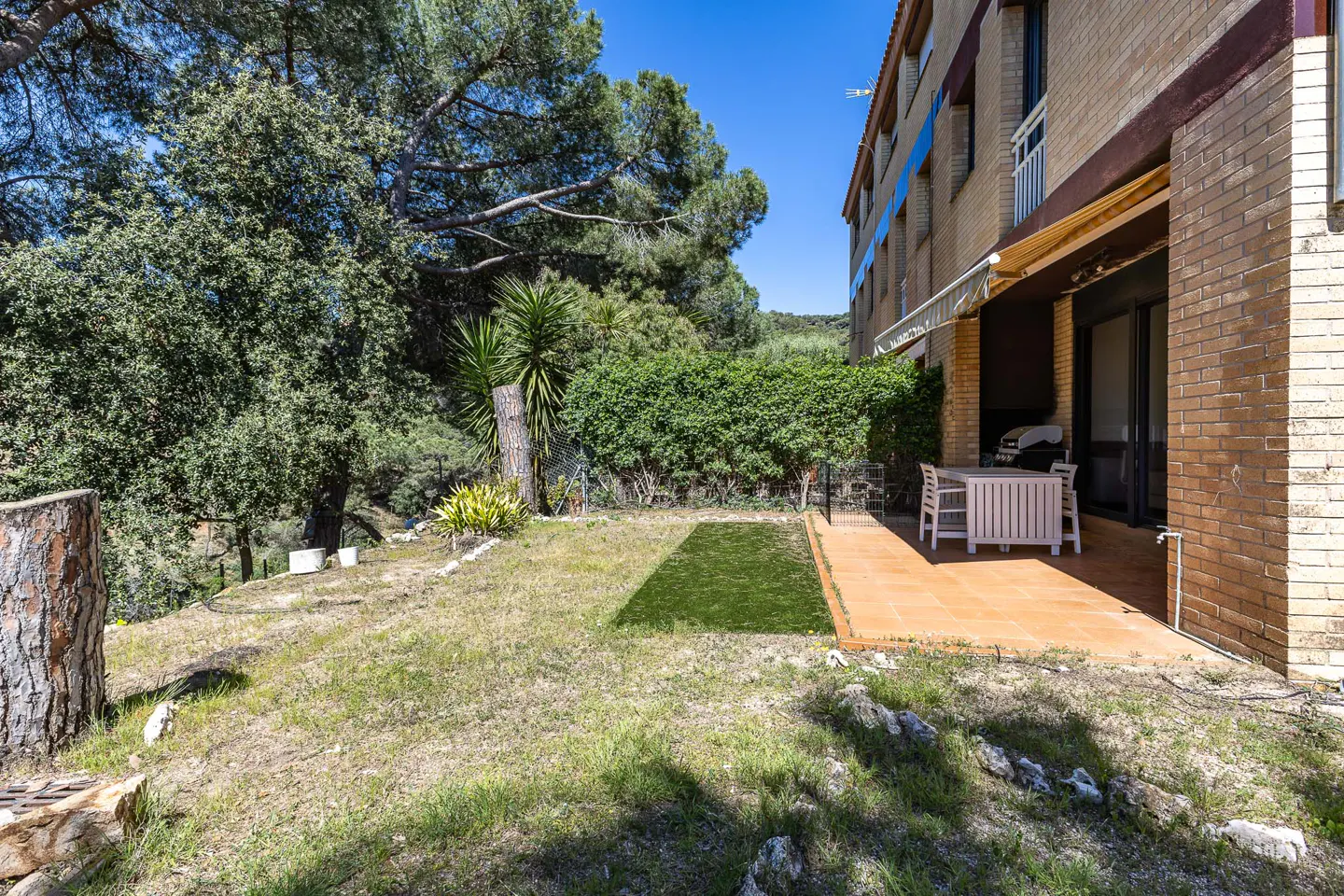 Brick building with a patio, table, and chairs. Green grass and trees surround the building. Blue sky.