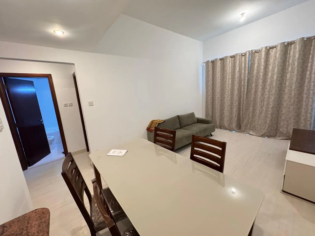 A bright, modern apartment interior with a dining table, sofa, and bathroom visible. The walls are white, and the curtains are beige.