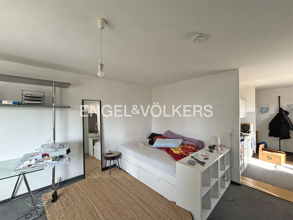 A bright studio apartment with a bed, desk, mirror, and a small kitchen area. The walls are white, and the floor is dark.