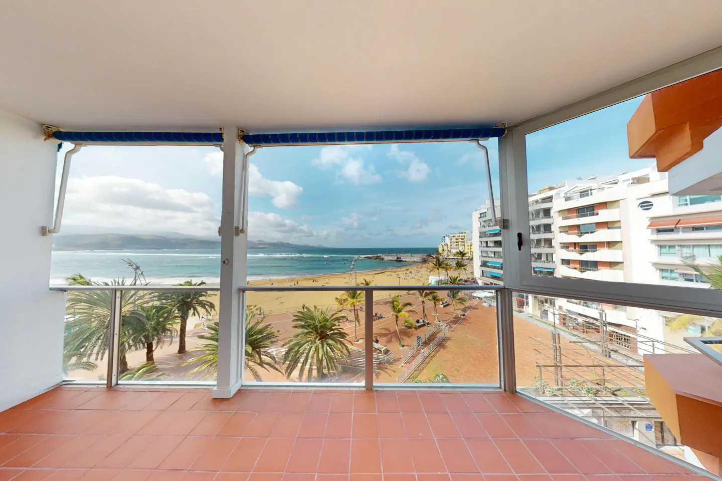 Balcony view of a beach with palm trees, sand, and ocean. Buildings are visible in the background. The balcony has red tile flooring.