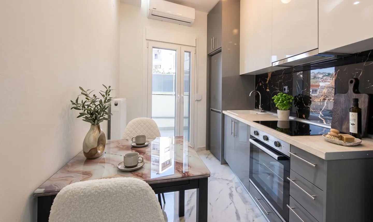 Modern kitchen with a marble table set for two, gray cabinets, and black marble backsplash. Olive branches in a vase add a touch of nature.
