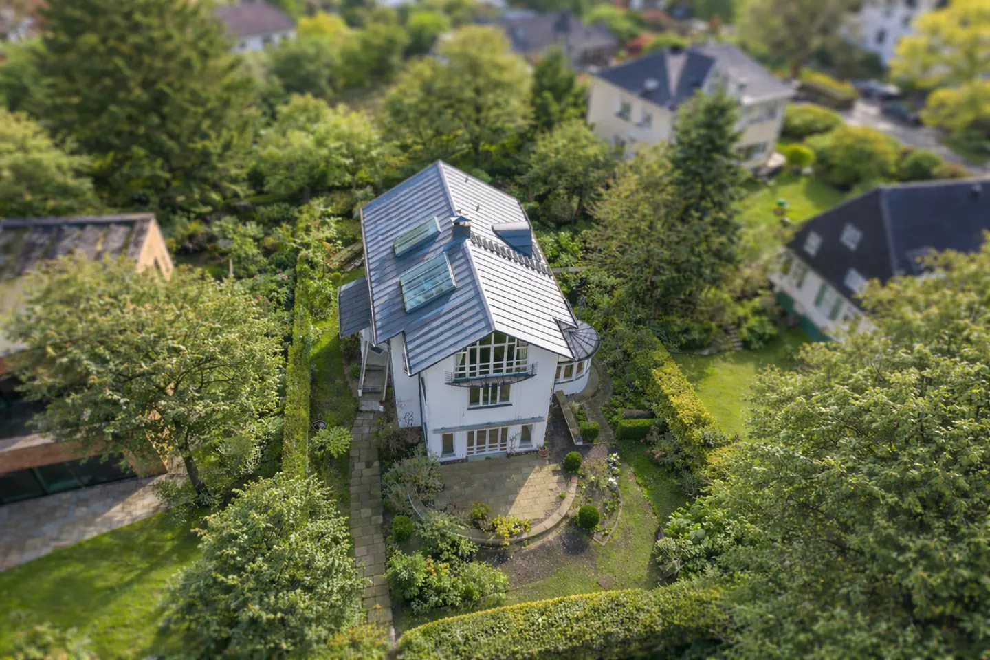 Aerial view of a white two-story house with a gray roof, surrounded by green trees and hedges. A stone path leads to a patio.