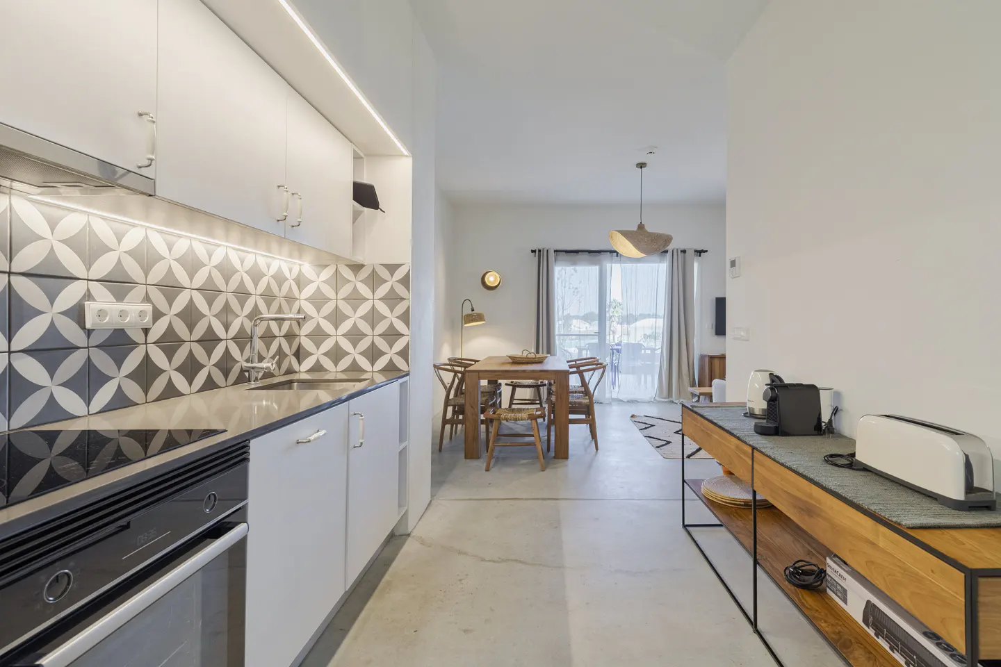 A bright, modern apartment interior with a kitchen, dining area, and living room. White cabinets, patterned backsplash, and wood furniture are visible.