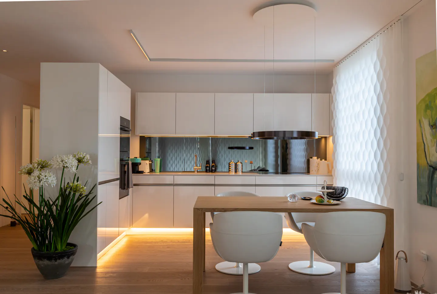 Bright, modern kitchen with white cabinets, wood floors, and a dining table with white chairs. A flower pot sits near the cabinets.
