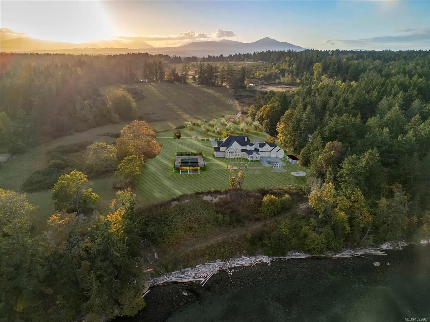 Aerial view of a large white house with a black roof, a tennis court, and a manicured lawn, surrounded by trees and water at sunset.