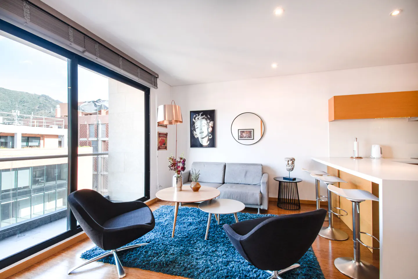 Modern living room with two black chairs, a grey sofa, blue rug, and a kitchen island with bar stools by a large window.
