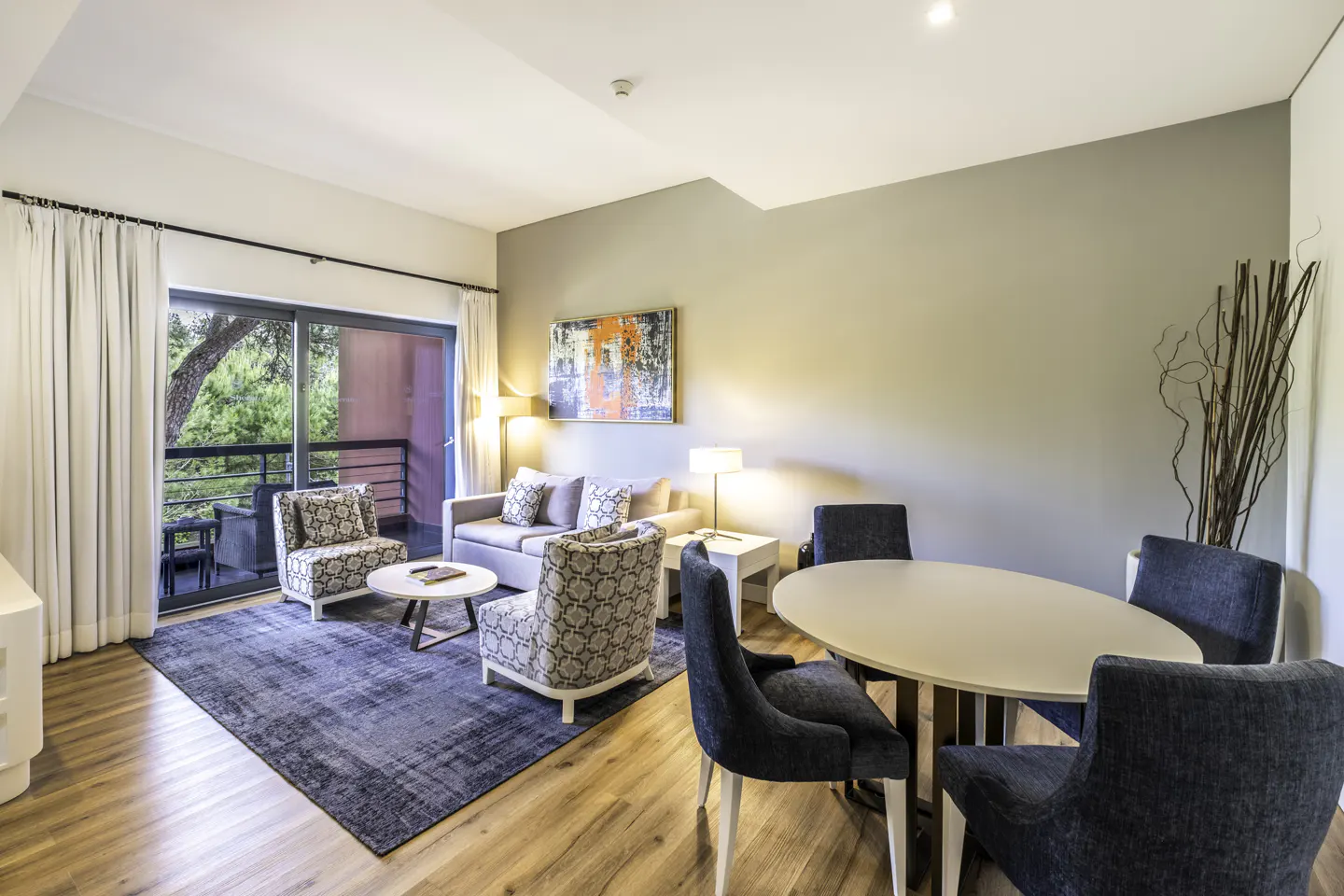 Living room with a sofa, two armchairs, a round table, and a dining table with four chairs. Balcony with trees in the background.