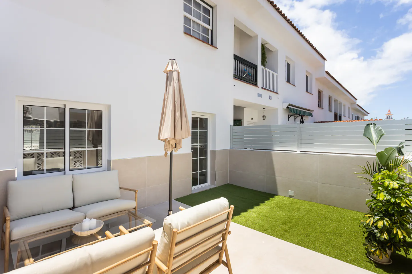 Outdoor patio with beige furniture, glass table, and closed umbrella. White building with windows and green lawn in the background.