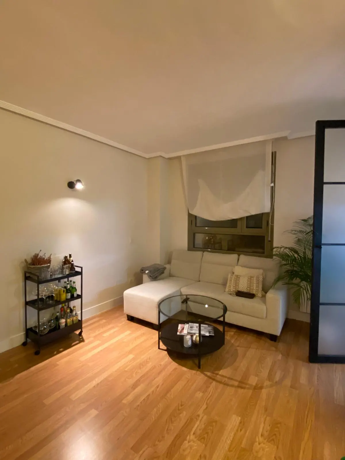 Modern living room with light wood floors, white sectional sofa, glass coffee table, and a black bar cart with drinks.