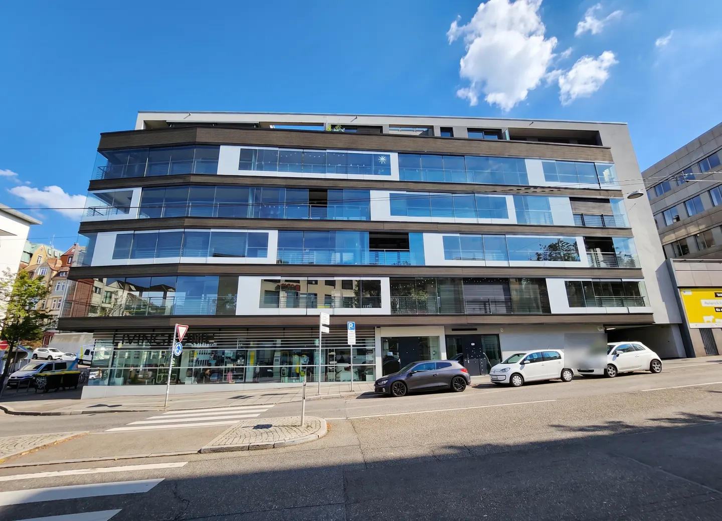 Modern apartment building with glass balconies and a retail space on the ground floor, cars parked on the street.