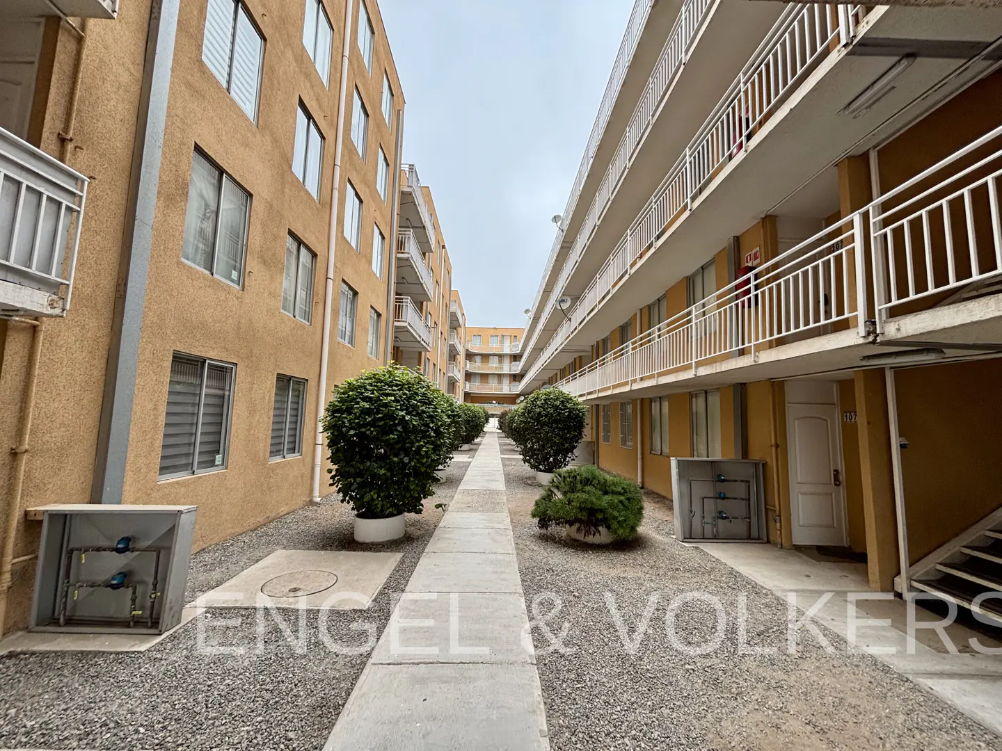 Exterior view of a multi-story apartment complex with a walkway, bushes, and gravel. The buildings are tan with white balconies.
