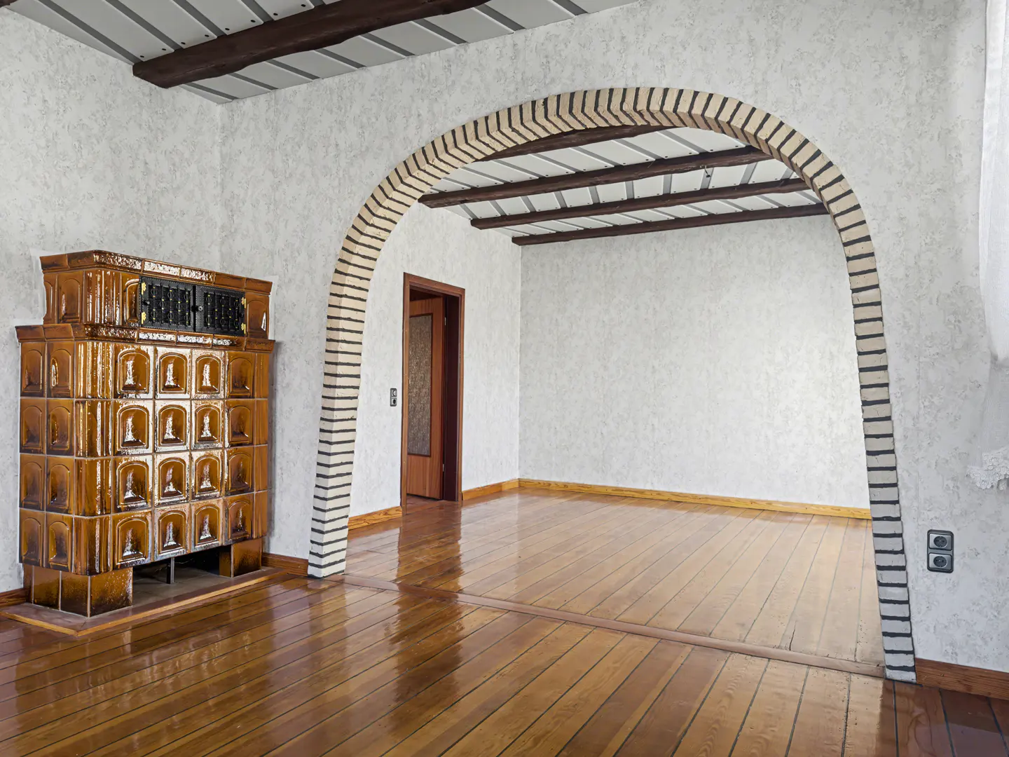 A room with a brick archway, wood floors, and a brown tiled stove.