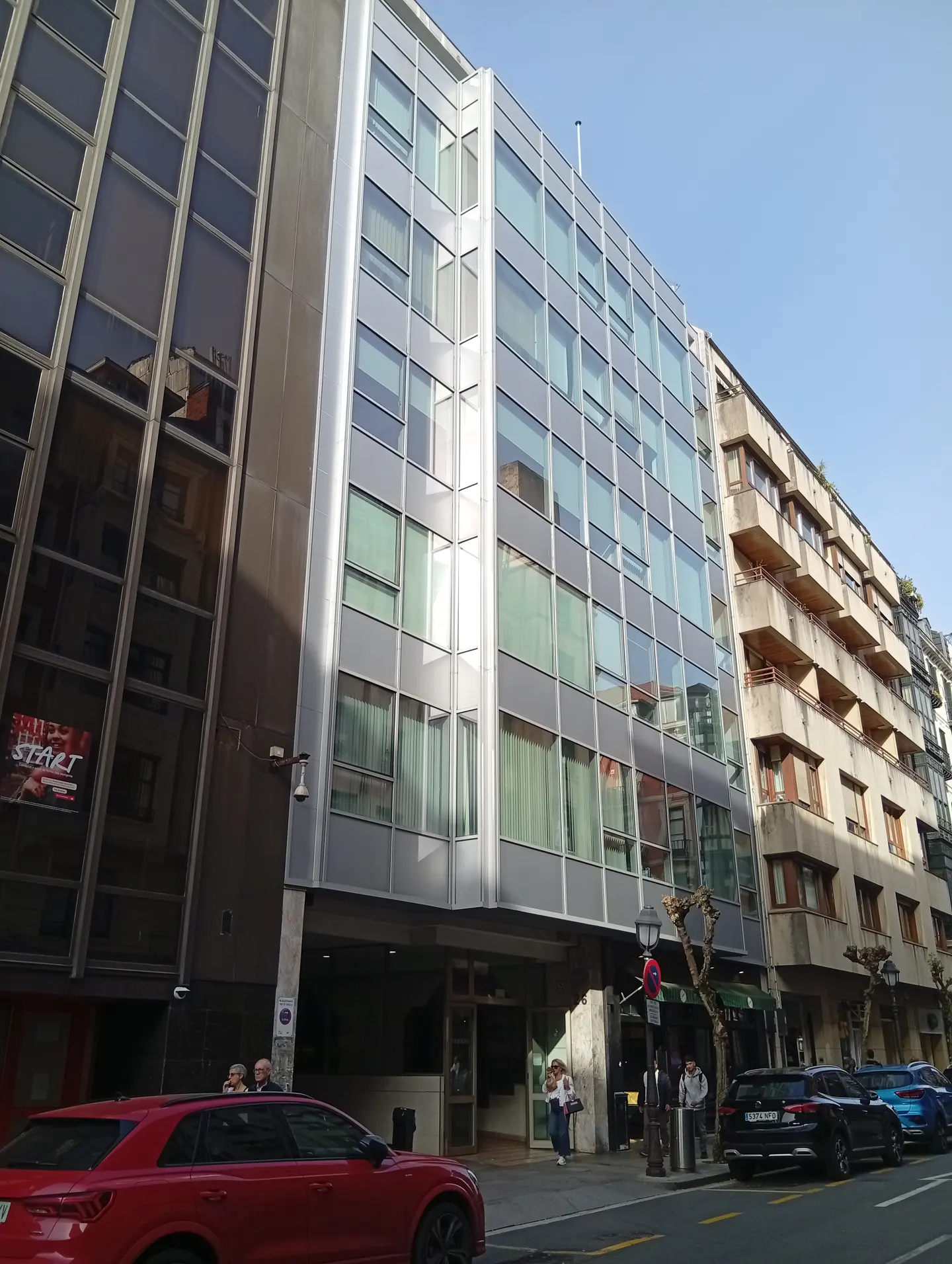 Street view of a modern, multi-story building with large windows, parked cars, and pedestrians on the sidewalk.