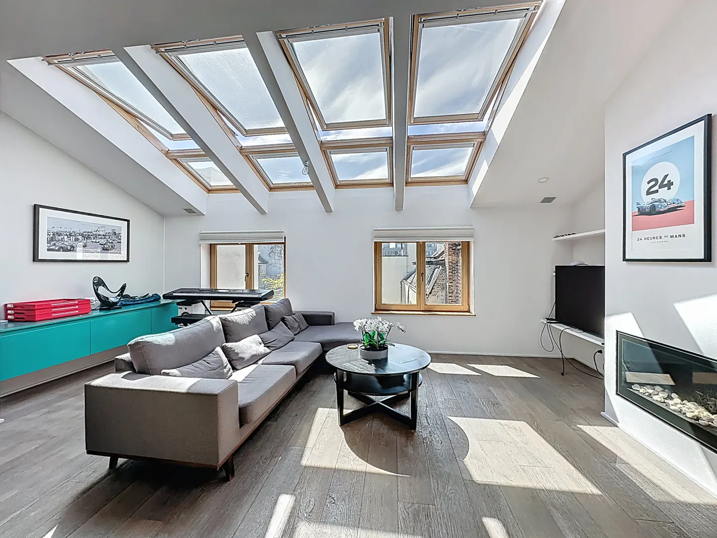 Bright living room with skylights, gray sectional sofa, round coffee table, and turquoise cabinet. Hardwood floors and white walls.