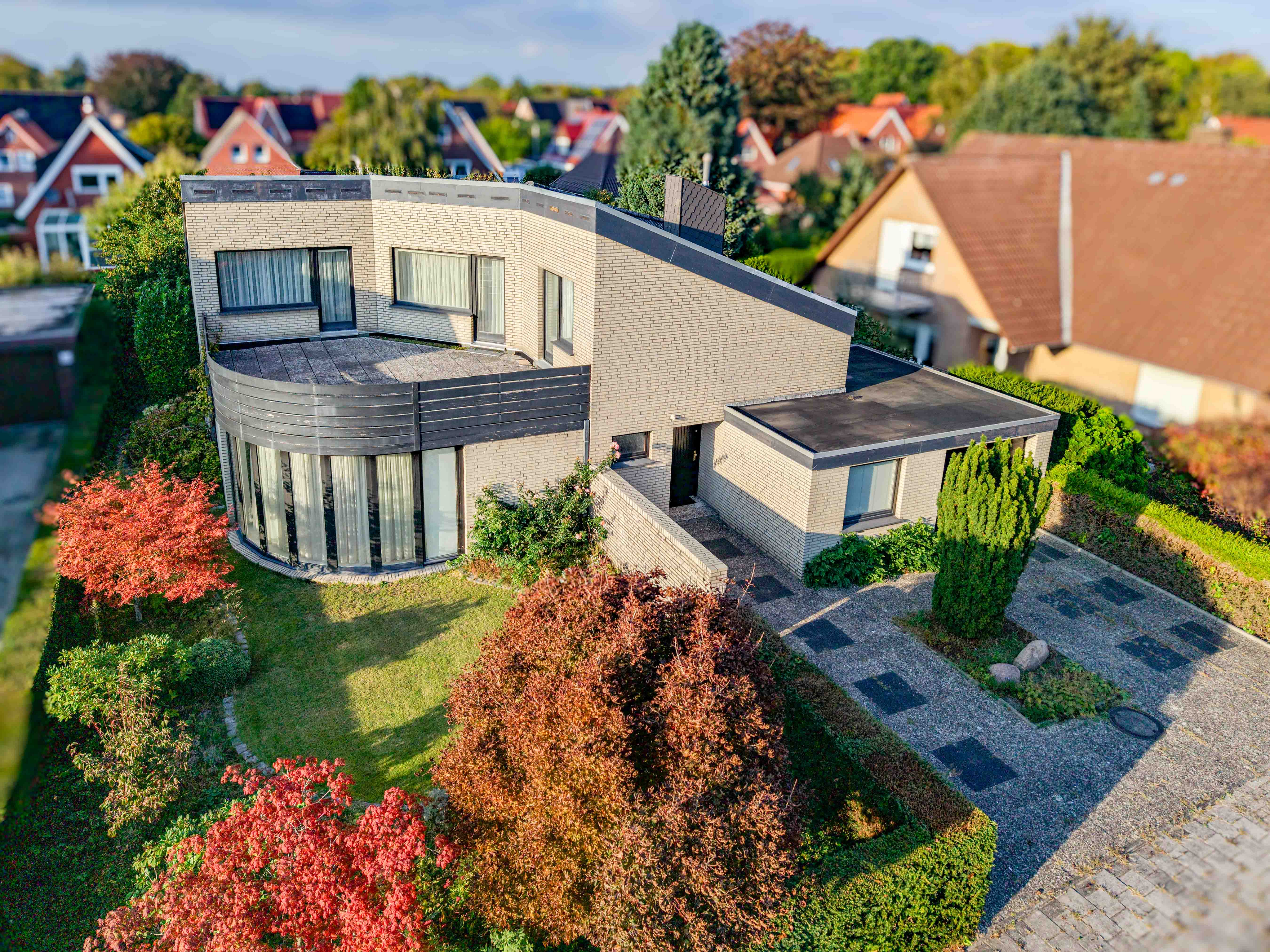 Aerial view of a modern beige brick house with a curved facade, flat roof, and autumn-colored trees in a suburban setting. [AI-generated text]