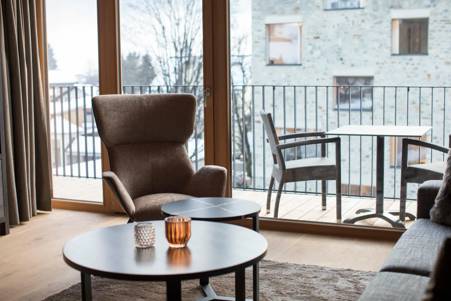 Modern living room with a brown armchair, two round coffee tables, and a balcony view of a stone building in winter.