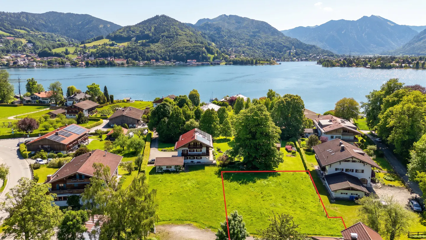 A vacant lot outlined in red, sits on a green hillside overlooking a blue lake and mountains. Houses and trees surround the property.