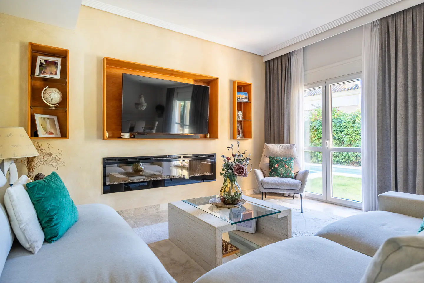Living room with a cream sofa, a glass coffee table, and a TV over a fireplace. Shelves flank the TV. A window with gray curtains is on the right.