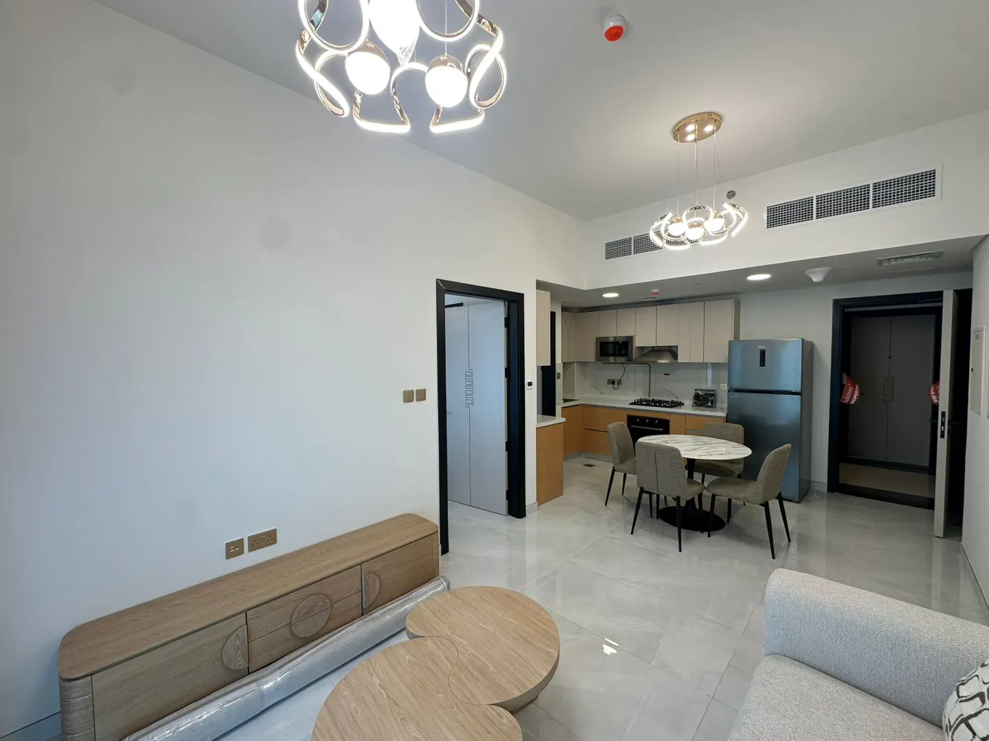 Bright, modern apartment interior with white walls, light wood furniture, and a round dining table. Kitchen with stainless steel appliances visible.