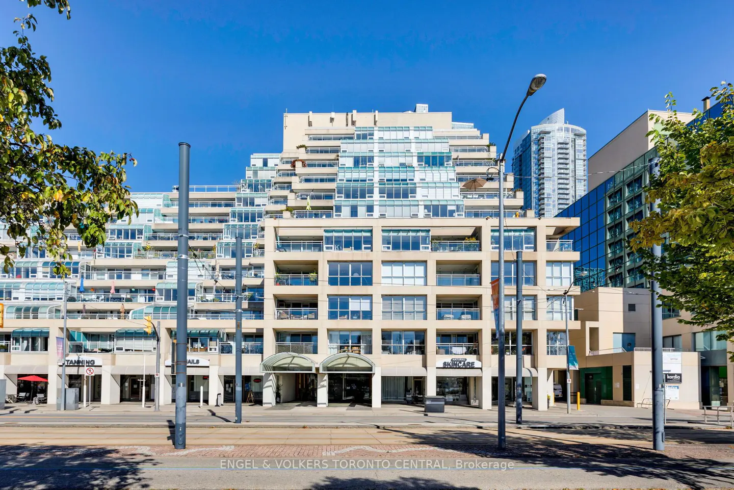 Exterior shot of a beige condo building with balconies and shops on the ground floor on a sunny day.