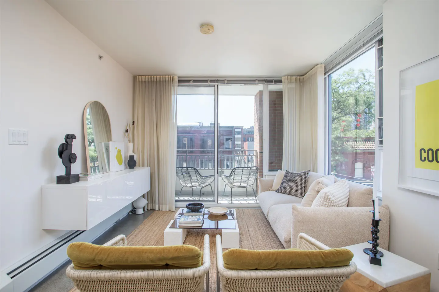 Bright living room with white walls, a beige sofa, and two wicker chairs with yellow cushions. A balcony is visible through sliding glass doors.