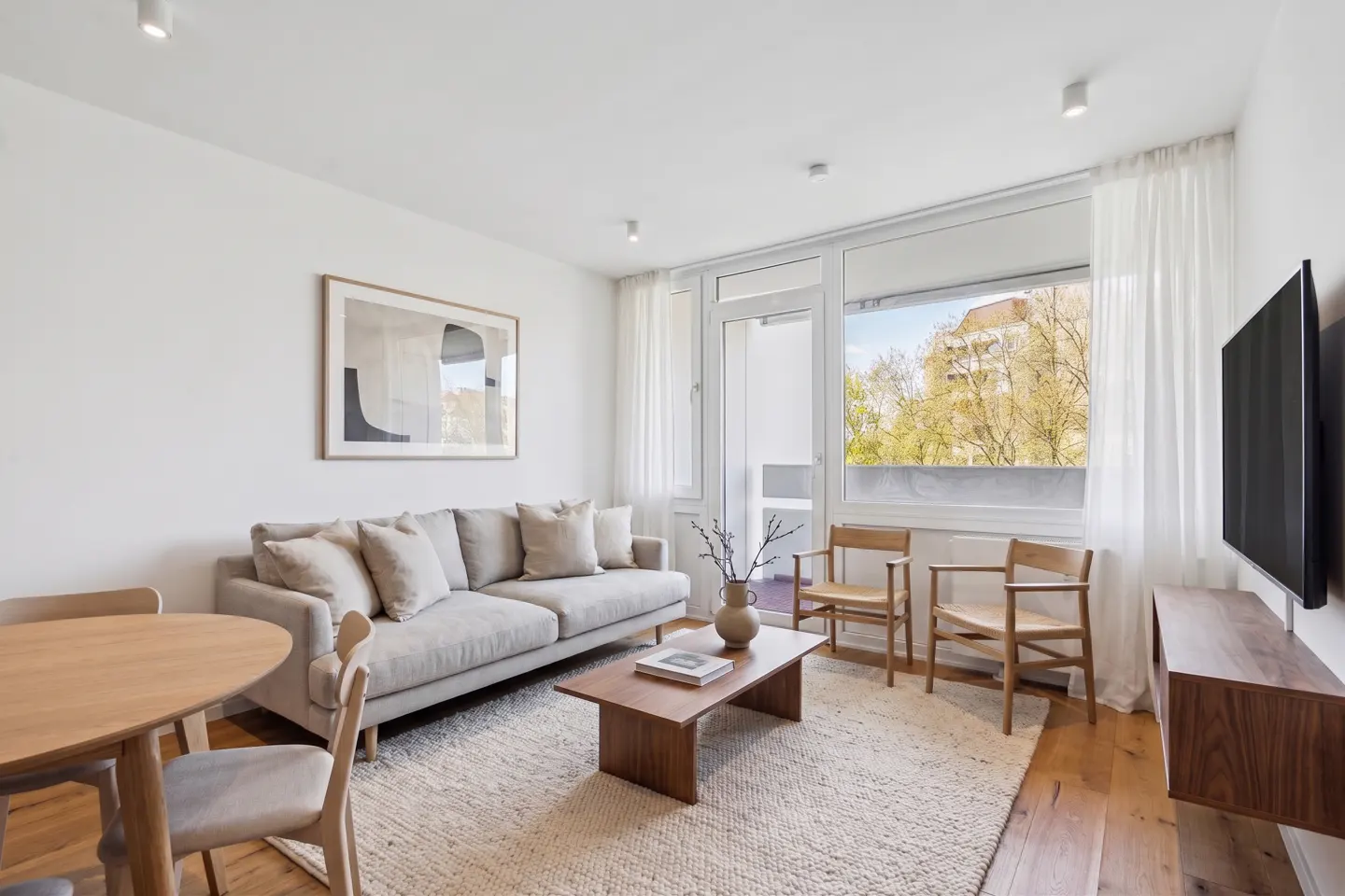 Bright living room with a beige sofa, wood table, chairs, and a large window with a view of trees.