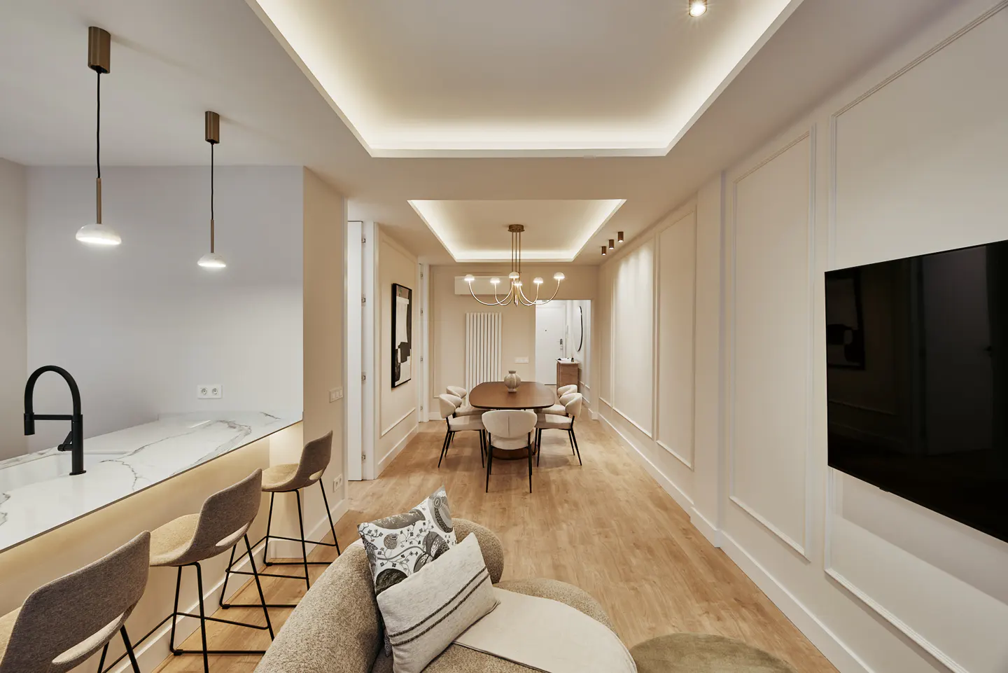 Interior view of a modern apartment with a kitchen island, dining table, and living area. Neutral tones, wood floors, and recessed lighting create a warm ambiance.