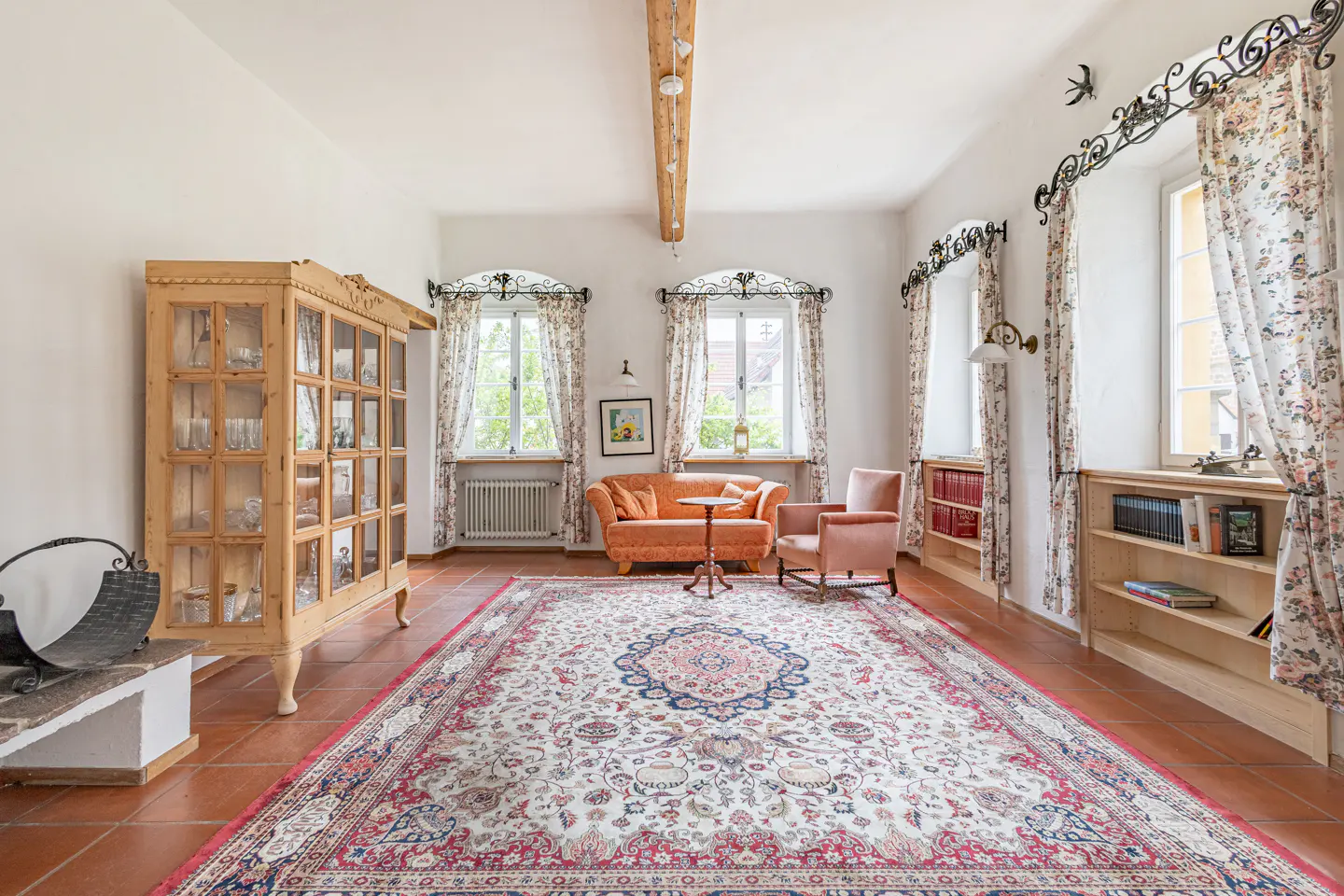 Bright living room with terracotta tile floor, large patterned rug, orange sofa and chair, and light wood cabinet.