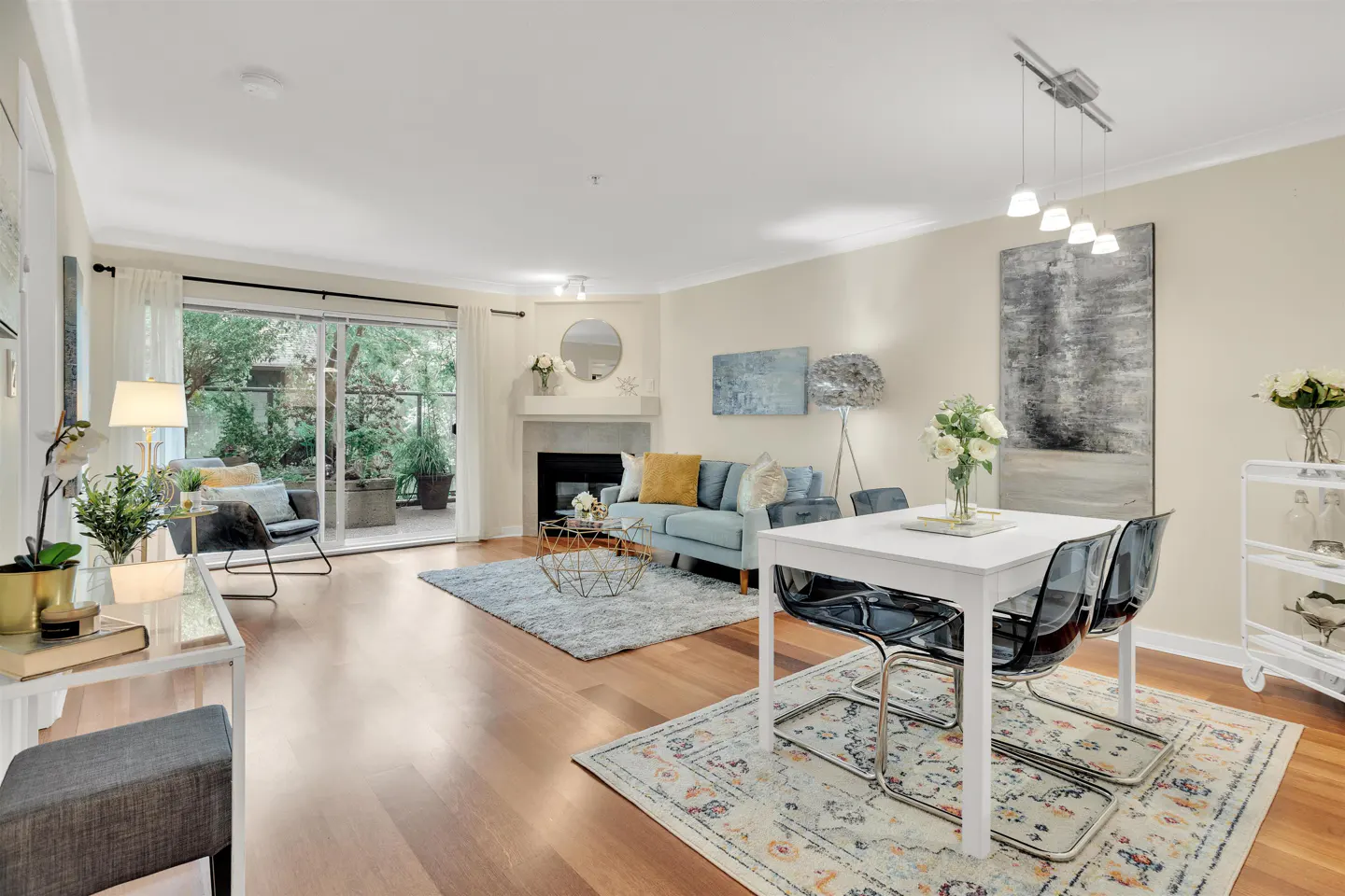 Bright living and dining area with hardwood floors, a blue sofa, fireplace, and a white dining table with transparent chairs.