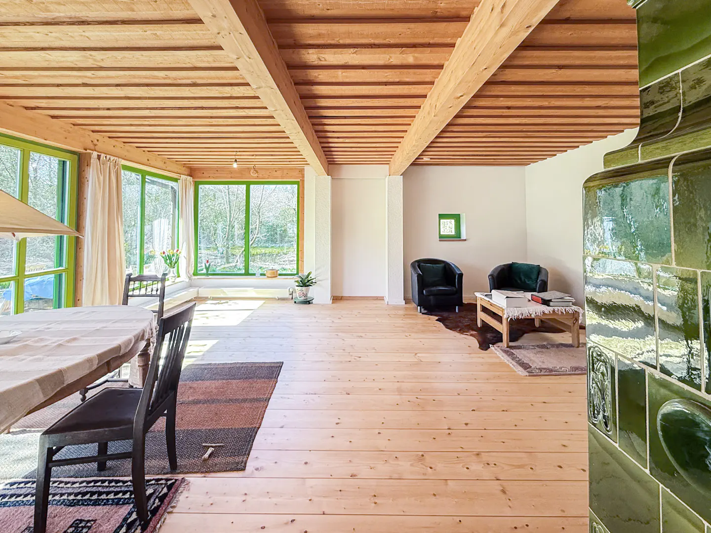 Bright room with wood floors, beams, and green window frames. A table and chairs are on the left, and armchairs and a coffee table are on the right.