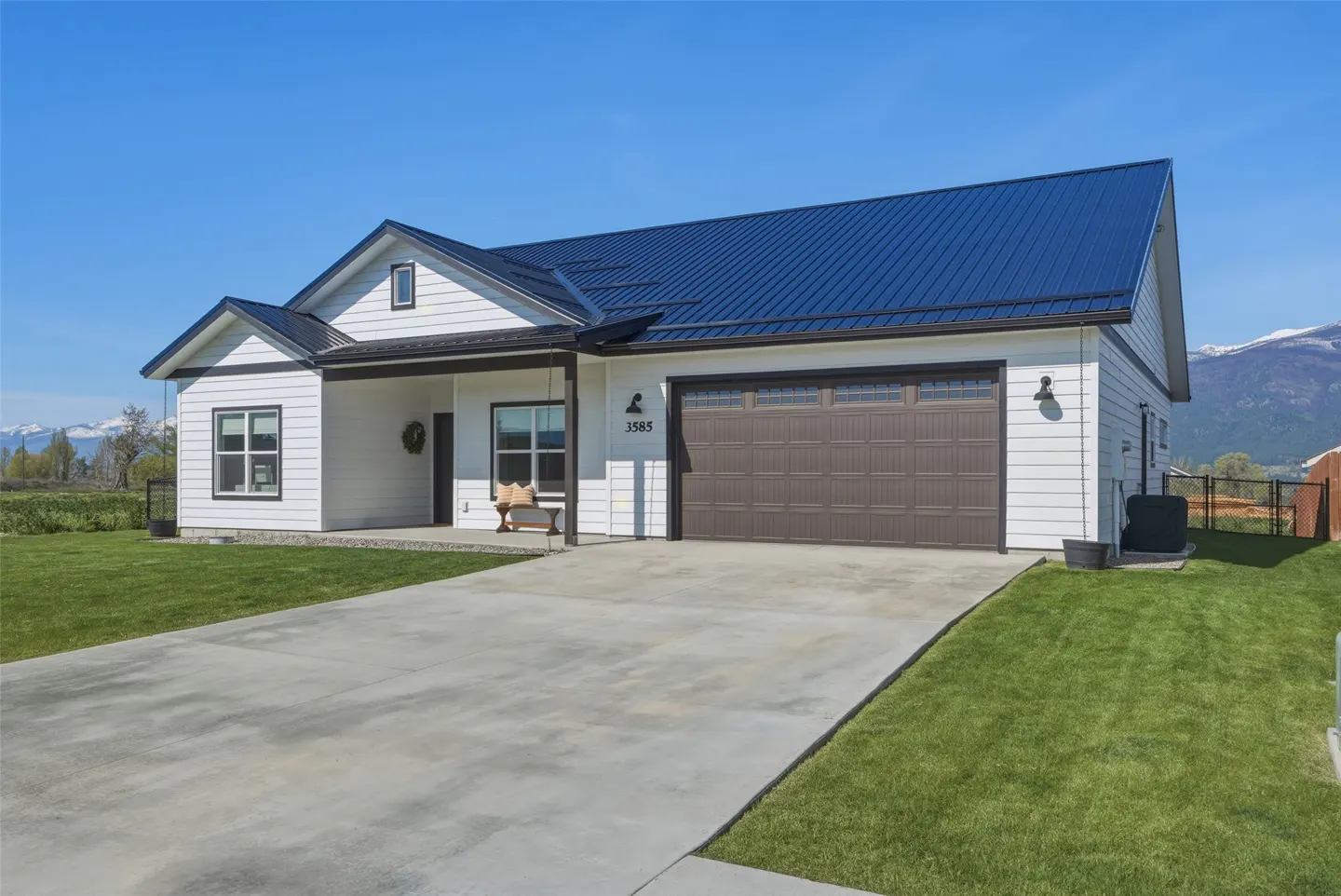 A white, one-story house with a blue metal roof, brown garage door, and a concrete driveway on a sunny day.