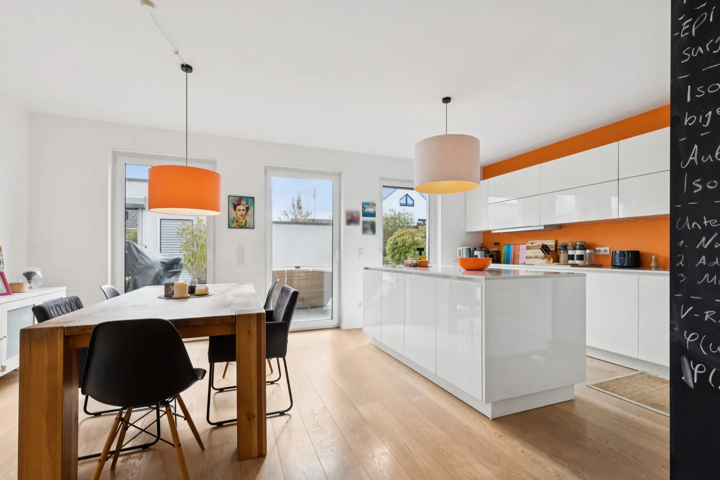 Bright, modern kitchen with wood floors, white cabinets, and orange accents. A wooden table with black chairs sits under an orange pendant light.