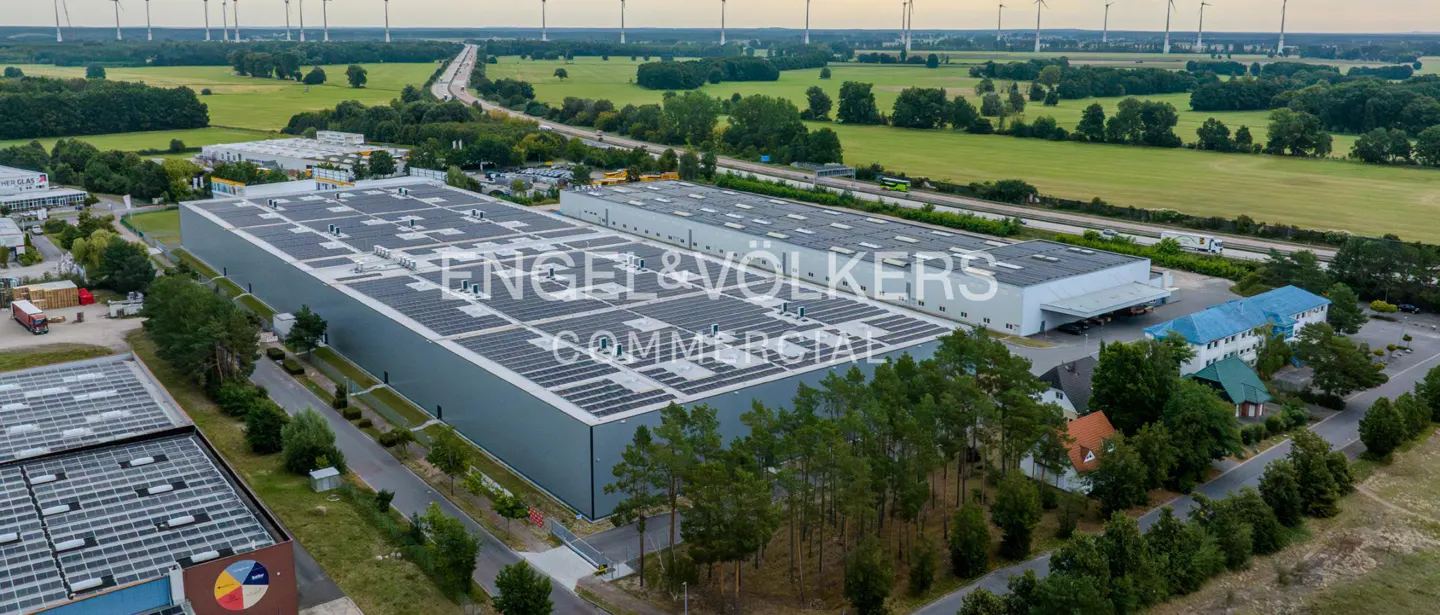 Aerial view of two large, gray commercial buildings with solar panels on the roof, surrounded by green trees and fields. Wind turbines are visible in the distance.