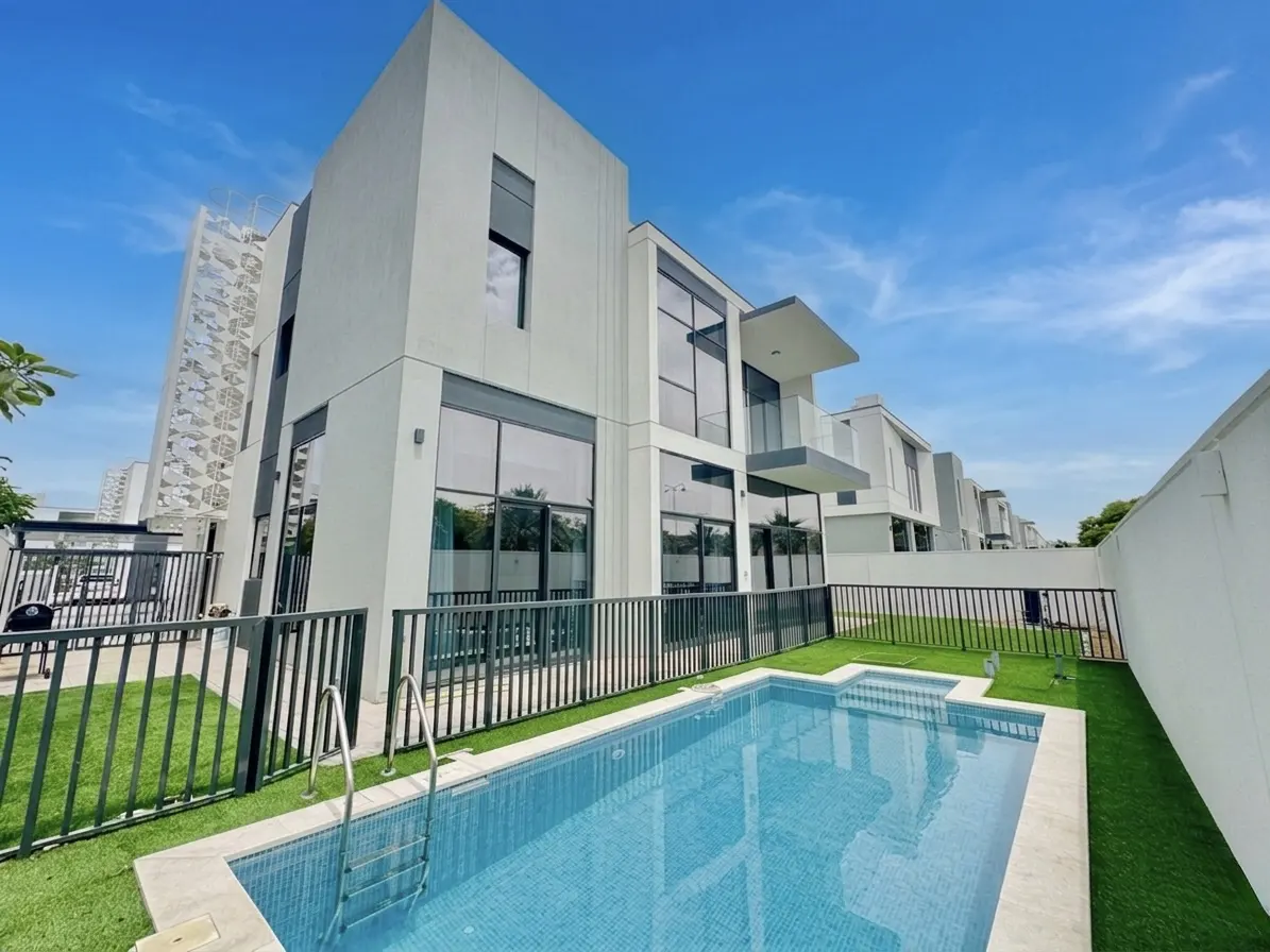 Modern white townhouses with a blue tiled pool in the foreground, surrounded by green grass and a black fence under a blue sky.