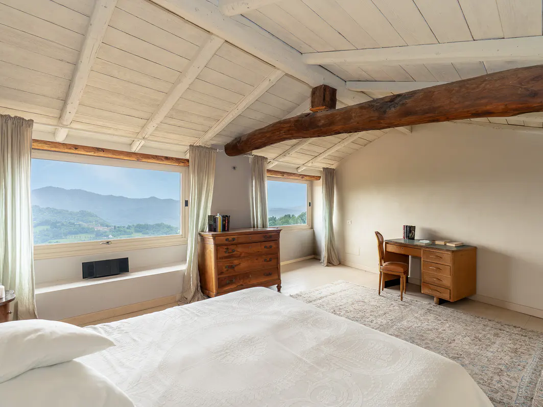 Bedroom with white bedding, wooden desk, and dresser. Windows overlook a green, hilly landscape. White, beamed ceiling with a rustic wooden beam.