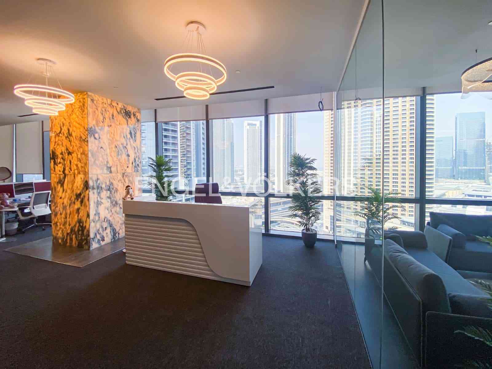 Office lobby with a white reception desk, marble accent wall, and city views through large windows. Modern, circular chandeliers hang from the ceiling. [AI-generated text]