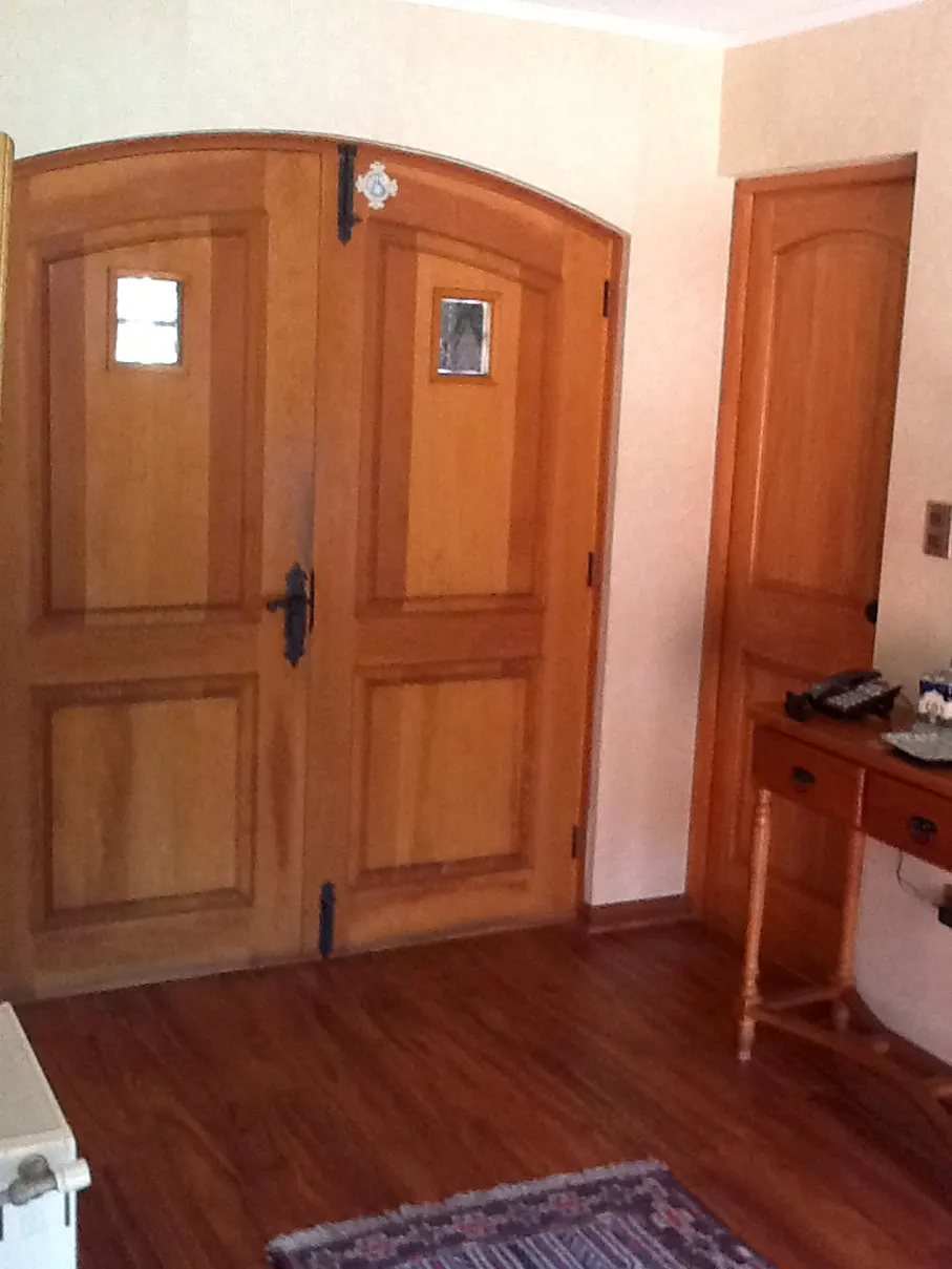 Interior view of a room with wood floors, a rug, and two wooden doors. One is a double door with small windows, the other is a single door. A wooden table is on the right.